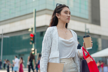A young asian female office employee walking at a busy public plaza in the city. Holding a chart, coffee and some shopping bags.