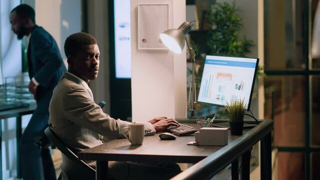 Portrait of african american computer operator sitting at desk during nightshift, looking through online financial business documents while coworker in background goes to bathroom