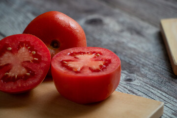 Un tomate a la mitad en una tabla de cortar. Todo se encuentra sobre una mesa gris rústica. Cerca...