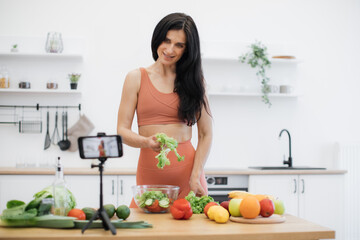 Cheerful caucasian adult adding leafy greens to salad bowl while looking at camera of cell phone in kitchen. Efficient food blogger starting online cooking course about healthy eating habits at home.