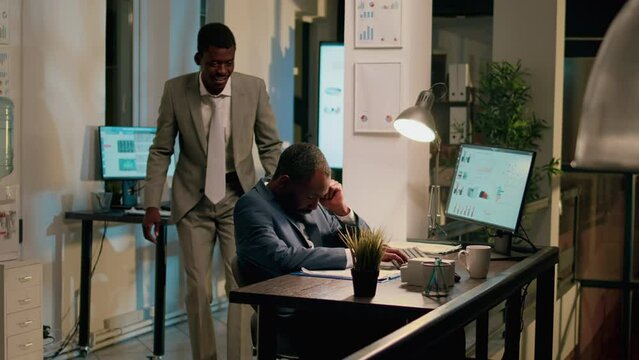 Employee playing practical joke on unsuspecting tired coworker asleep in office during nightshift. Fatigued businessman sleeping overnight at work, being woken up by colleague
