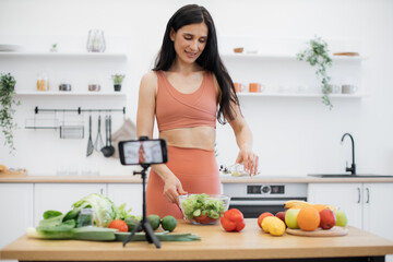 Beautiful young woman in sportswear adding oil to vegan salad on webcam of smartphone fixed by tripod in kitchen. Fit and healthy adult recommending fresh ingredients for health blog subscribers.