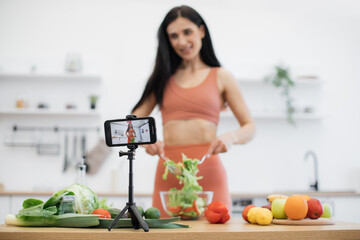 Digital phone on tripod placed on kitchen table among groceries shooting smiling lady preparing vegan salad. Cheerful food influencer covering nutrition related concern on online blogging.