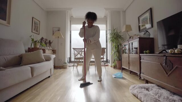 Young African American girl dancing and singing with broom doing housework living room. Black people sweeping the floor with brush and dustpan to clean house. Cheerful woman doing domestic life.