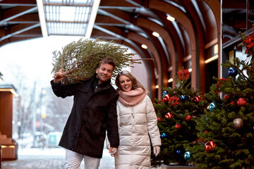 Beautiful young couple in love buying a Christmas tree from the market to decorate their home. Caucasian man and woman are preparing for Christmas. Anticipation of the New Year Eve in a snowy city.