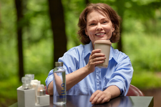 Adorable Mature Woman Sits At The Table In Outside Cafe With Paper Coffee Cup And Bottle Of Water. Middle-aged Brunette In Blue Shirt Smiling Joyfully. Senior Lady Enjoying Life On Summer Terrace.