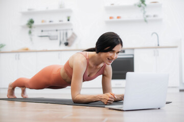 Side view of caucasian fitness woman dressed in activewear doing plank exercise on yoga rug while staying at home. Fit lady listening motivational tracks on remote laptop. Concept of active lifestyle.