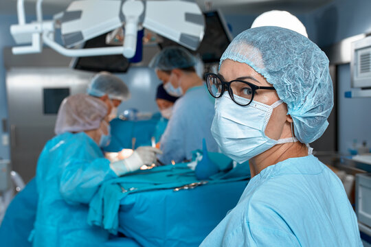 Close-up portrait of a female doctor in glasses and face mask on the background of surgeons team during complex surgical operation in a sterile operating room. Modern medicine, saving patients life.