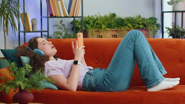 Portrait of happy overjoyed woman in wireless headphones relaxing lying on couch at home apartment choosing listening favorite energetic disco dancing music. People weekend daytime leisure activities