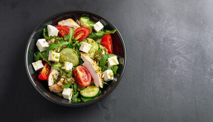 Tasty appetizing fresh salad with chicken, tomatoes, cucumbers, pesto sauce and cheese feta in bowl on black background. View from above. Horizontal with copy space.