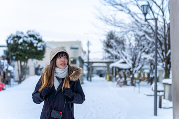 Fototapeta na wymiar Young Asian woman in winter coat walking on city street covered in snow in snowy day evening. Attractive girl enjoy urban outdoor lifestyle travel in Japan on holiday vacation in winter season.