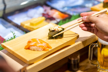 Naklejka na ściany i meble Asian woman hand using chopsticks eating Japanese food nigiri sushi with seaweed on sushi board on open kitchen counter. Chef serving assorted sushi and sashimi to customer at Japanese restaurant.