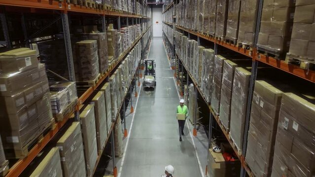 Worker Operating Hand Pallet Truck, Moving Cardboard Boxes Between Shelves Filled with Goods in Warehouse. People Work in Product Distribution Logistics Center Inside