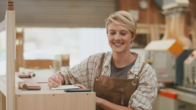 Portrait of smiling female apprentice carpenter in furniture workshop writing in notebook - shot in slow motion