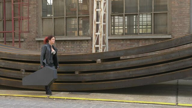Rocker Girl carrying a coffin guitar case through an Industrial place, Full shot slow motion.