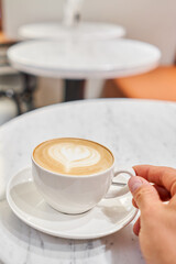A man holds a cup with his hand A cup of hot cappuccino on the table in the coworking space of the coffee shop. Coffee break at the cafe. Close-up