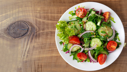 Salad with greens, cherry tomatoes, red onions and cucumber on white plate with wood background, overhead view