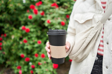 A cup of hot drink in the hands of a young woman. A girl in the park holds a cup of coffee in her hands. Breakfast on the go. The concept of a picnic and going out.