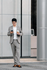 Asian businessman holding a cup of coffee is using mobile phone outside the modern office building. 