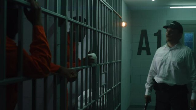 Warden with baton comes to jail cells and starts bullying prisoners. African American men serve imprisonment term in correctional facility or detention center. Guilty murderers in gloomy prison cells.