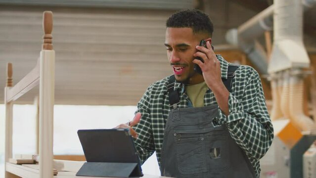 Male apprentice carpenter in furniture workshop using digital tablet and talking on mobile phone - shot in slow motion