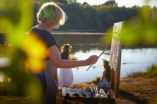 Adult female artist painting picture near water of river or lake in nature and girls in white sundress and flower wreath posing in the holiday of Ivan Kupala in nature at sunset