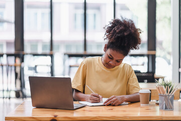 Obraz premium Young African American woman is taking notes in a notebook while watching a webinar video course. Afro american student listening to the lecture to study online through e-learning.