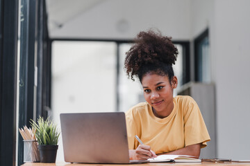 An attractive and happy millennial businesswoman is sitting on a sofa in her living room, working with a laptop computer at home.