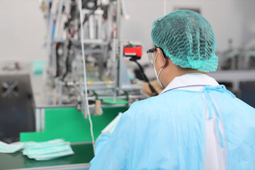 Rear view worker in personal protective equipment or PPE inspecting quality of mask and medical face mask production line in factory, manufacturing industry and factory concept.