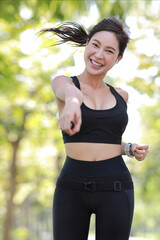 Beautiful asian fitness female sportswoman runner doing morning jogging exercise training outdoor. Happy smile athlete woman running under the tree in the park while looking at camera and listen music