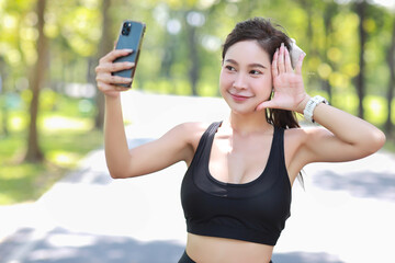 Portrait of happy and relax young asian sportswoman take a break after morning workout. Beautiful athlete smiling woman doing selfie by using mobile after training run in the park near lake and trees