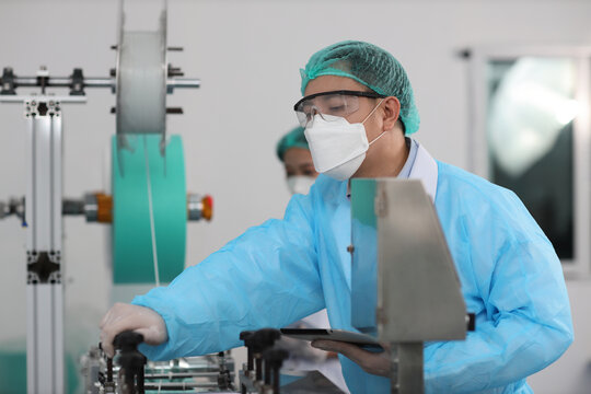Worker in personal protective equipment or PPE inspecting quality of mask and medical face mask production line in factory, manufacturing industry and factory concept.