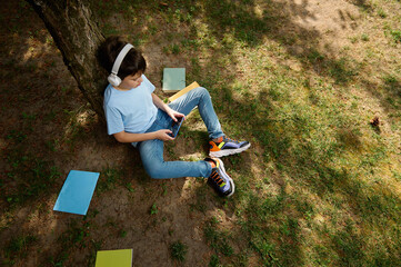 Fototapeta premium Schoolboy in wireless headphones, top view, playing online video games. Kids Education Digital addiction Entertainment