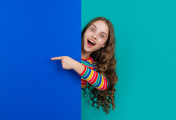 surprised teen child behind blank blue paper with copy space for advertisement. point finger