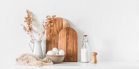 Cozy scandinavian kitchen interior background - dishes cutting boards,  bowl with eggs, a jug of dried flowers on a white table