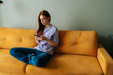 Portrait of frustrated young woman sitting on yellow couch looking at smartphone screen thinking about problem difficulty, feeling failure and exhausted, suffering grief and bad relationship