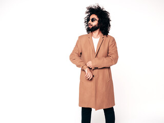 Handsome smiling hipster model.  Unshaven Arabian man dressed in brown coat clothes. Fashion male with long curly hairstyle isolated on white in studio. Cheerful and happy. In sunglasses