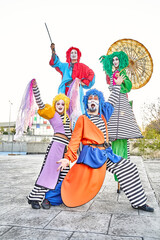 Group of face-painted performers in colorful clown costumes and wigs juggling and dancing on stilts...