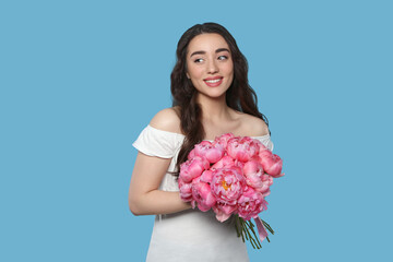 Beautiful young woman with bouquet of pink peonies on light blue background
