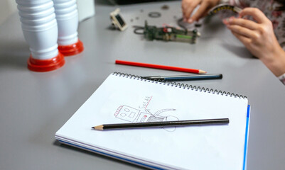 Robot draw by child on notebook with note about solar panel while unrecognizable hands of student...