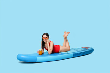 Happy woman with refreshing drink resting on SUP board against light blue background