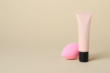 Tube of skin foundation and sponge on beige background, space for text. Makeup product