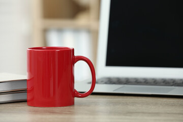Red ceramic mug, notebooks and laptop on wooden table indoors. Space for text