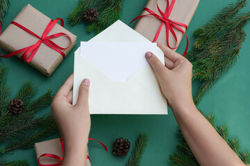 Woman hands holding blank paper and envelope . Flat lay of green background with gift boxes and Christmas decoration. Top view mock up and copy space for text.