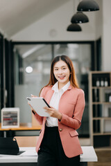 Confident Asian woman with a smile standing holding notepad and tablet at the office..