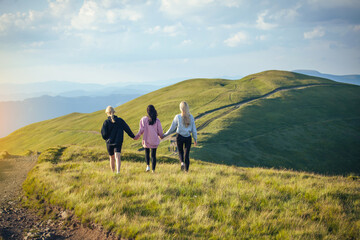 Fototapeta na wymiar Three Young Girl Friends Walk Together Holding Hands into Beautiful Mountain Landscapes