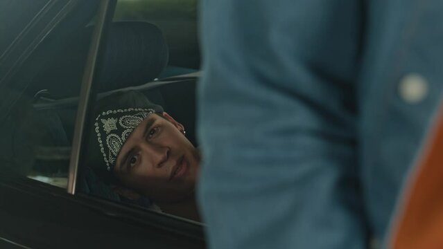 Caucasian cool guy in white undershirt and bandana on his head showing cash rolls at camera while rapping in backseat of black retro car