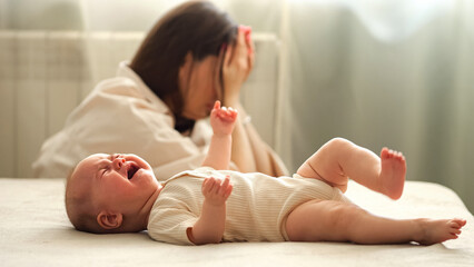 Tired infant girl cries on double bed against depressed mother sitting on floor. Baby daughter...