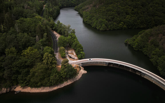 Aerial view of road with green woods by lakes water and mountains in summer Luxembourg. Summer road trip on white car across the river dam
