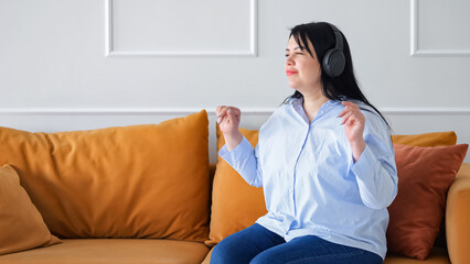 Fototapeta na wymiar Adult woman sitting on sofa listens to favourite music. Black-haired lady wearing headphones enjoys listening to energetic songs at home
